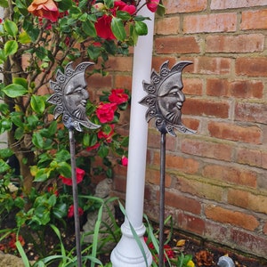 May include: Two black metal garden stakes featuring a sun and moon design. The stakes are topped with a stylised face and have a rustic finish.