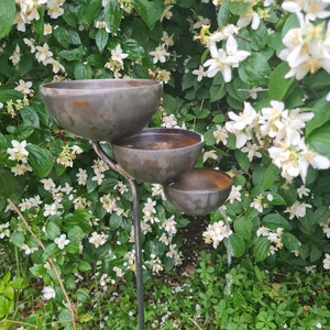 Puede incluir: Un comedero para p&aacute;jaros de metal con tres niveles de cuencos. El comedero est&aacute; situado en un jard&iacute;n con flores blancas y follaje verde.