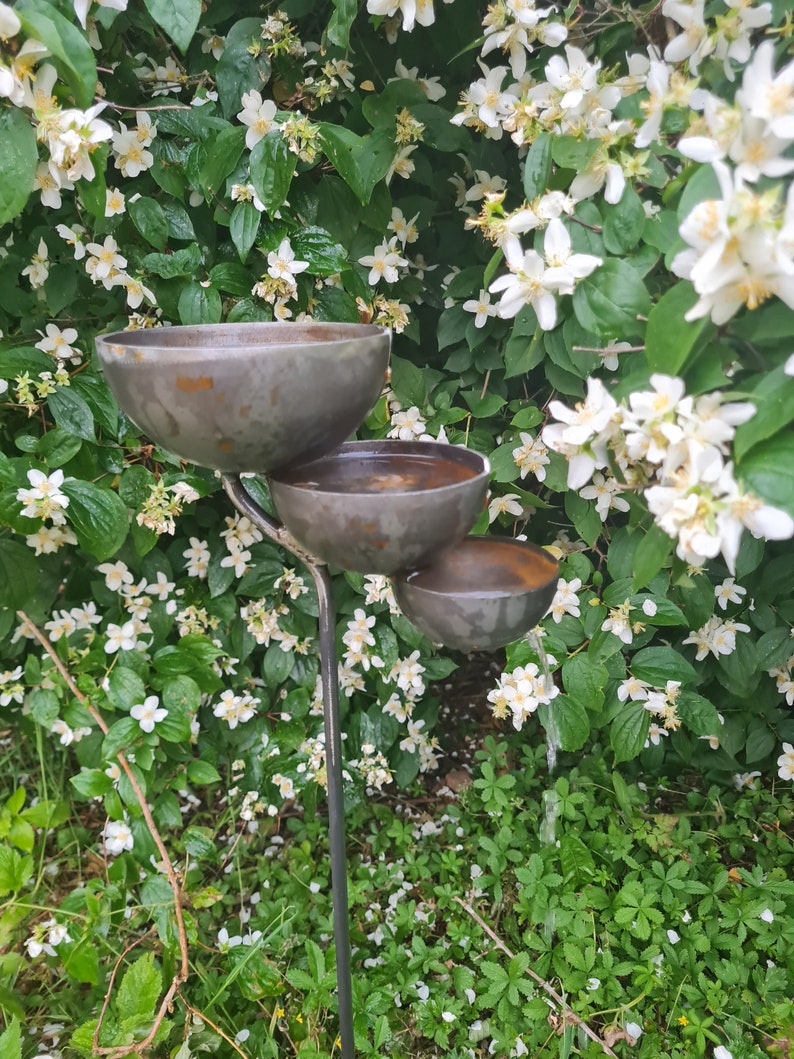 Puede incluir: Un comedero para p&aacute;jaros de metal con tres niveles de cuencos. El comedero est&aacute; situado en un jard&iacute;n con flores blancas y follaje verde.