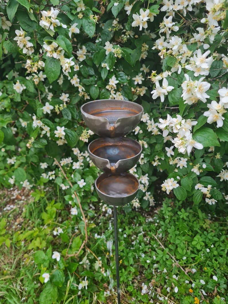 Puede incluir: Una fuente de agua de metal de tres niveles con un acabado r&uacute;stico. La fuente est&aacute; hecha de tres cuencos apilados uno encima del otro, con agua que fluye del cuenco superior al cuenco inferior. La fuente est&aacute; rodeada de vegetaci&oacute;n verde y flores blancas.