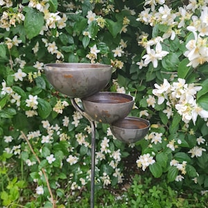 Puede incluir: Un comedero para p&aacute;jaros de metal con tres cuencos apilados en un poste alto de metal. El comedero est&aacute; rodeado de flores blancas y follaje verde.