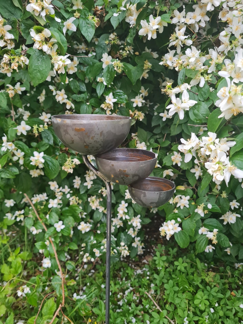 Puede incluir: Un comedero para p&aacute;jaros de metal con tres cuencos apilados en un poste alto de metal. El comedero est&aacute; rodeado de flores blancas y follaje verde.