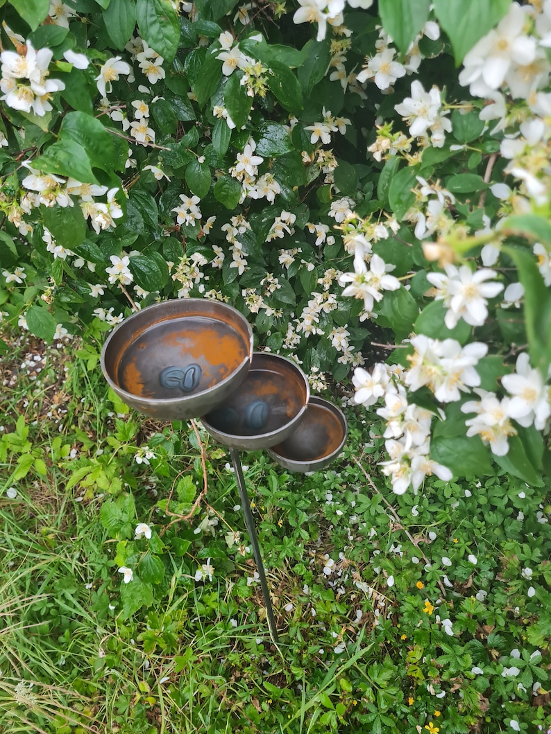 Puede incluir: Un bebedero para p&aacute;jaros de metal de tres niveles con un acabado marr&oacute;n oxidado. El bebedero para p&aacute;jaros est&aacute; situado en un jard&iacute;n con hierba verde y flores blancas.
