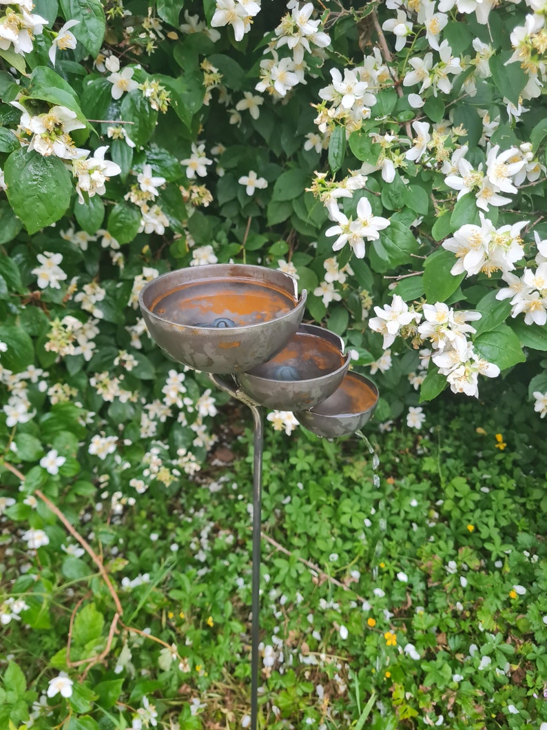 Puede incluir: Una fuente de jard&iacute;n de metal con tres niveles de cuencos. Los cuencos son de color marr&oacute;n oxidado y el agua fluye del cuenco superior al inferior. La fuente est&aacute; rodeada de hierba verde y flores blancas.