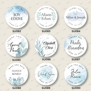 Op de afbeelding: Negen ronde blauwe en witte stickers voor een huwelijksdatum met aquarelontwerpen en kalligrafietekst. De tekst bevat de namen van de stellen en de datum van de bruiloft. De stickers zijn gelabeld met de productcode SU082 tot en met SU090.