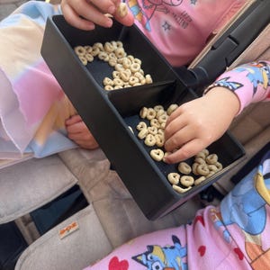 Könnte beinhalten: Eine schwarze, geteilte Schale mit Müsli gefüllt. Kinderhände greifen in die Schale, um das Müsli zu greifen. Die Kinder tragen rosa Pyjamas und Hemden mit Zeichentrickfiguren.