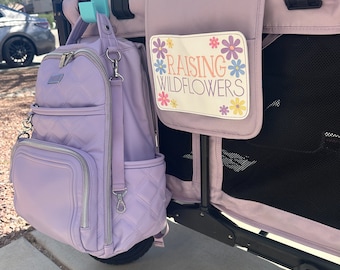 May include: A lavender quilted backpack with a silver zipper and a silver D-ring hanging from the strap. The backpack is hanging on a stroller with a pink and white floral canopy. The stroller has a black mesh side panel and a white patch with the text "Raising Wildflowers" in orange and pink.