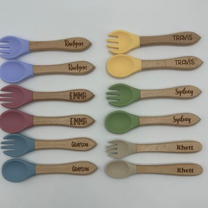 Może przedstawiać: Zestaw dziesięciu spersonalizowanych łyżeczek i widelczyków dla niemowląt z drewnianymi uchwytami. Łyżeczki i widelczyki są w różnych kolorach, w tym niebieskim, zielonym, żółtym i różowym. Każdy uchwyt jest grawerowany innym imieniem, w tym Travis, Sydney, Emma, Grayson i Raelynn.