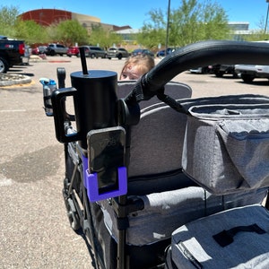 May include: A black stroller with a grey storage bag, a black cup holder with a black cup and a straw, and a purple phone holder with a black phone displaying the time 12:08.