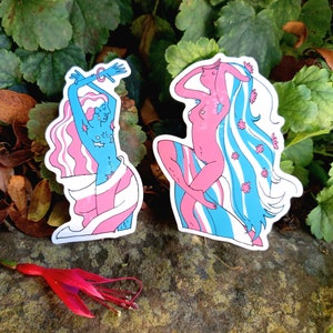 May include: Two colourful stickers featuring stylised figures of women with long hair. The figures are depicted with pink and blue stripes and flowers. The stickers are on a brown, textured surface.
