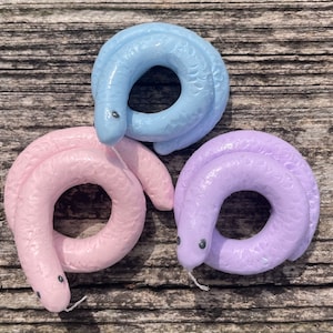 May include: Three ceramic rings in pastel colors: light blue, pink, and lavender. Each ring is shaped like a curled snake with a small black eye detail.