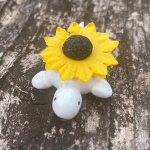 May include: A white ceramic turtle figurine with a yellow sunflower on its back. The turtle has black eyes and a small mouth.
