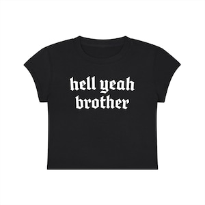 Puede incluir: Un top corto negro de manga corta con el texto blanco "hell yeah brother" impreso en una fuente de estilo gótico en negrita en el centro del pecho. La prenda tiene un cuello redondo y un dobladillo recto, mostrando un diseño sencillo.