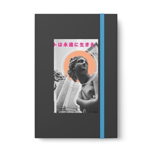 May include: Black notebook with a blue elastic band closure. The cover features a black and white image of a classical statue with a peach-coloured circle and Japanese text. The notebook has a minimalist design.