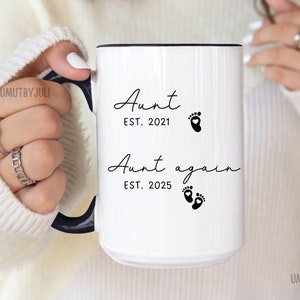 Könnte beinhalten: Weiße Keramiktasse mit schwarzem Text, der "Aunt EST. 2021" und "Aunt again EST. 2025" lautet, mit einer Fußabdruckgrafik neben jeder Textzeile.
