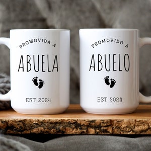 以下が含まれることがあります： 黒い文字と足跡のデザインが施された白いセラミック製のマグカップ2つ。左側のマグカップには「Promovida a Abuela Est 2024」と、右側のマグカップには「Promovido a Abuelo Est 2024」と書かれています。