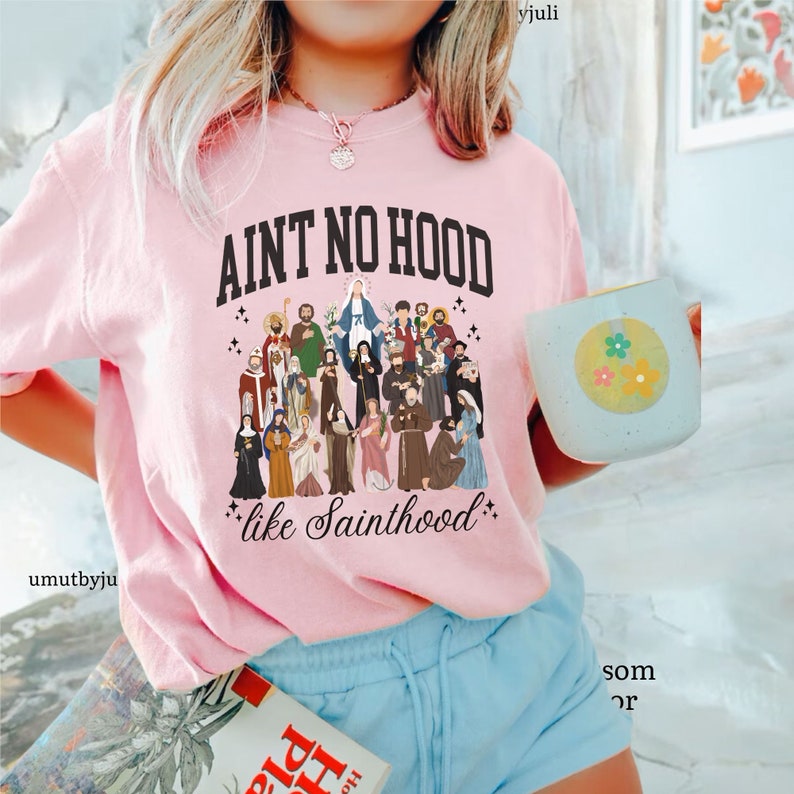 May include: Pink t-shirt with the text "Ain't no hood like Sainthood" and a graphic of a group of saints.