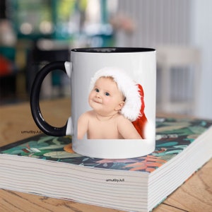 May include: White ceramic mug with a black handle. The mug features a printed image of a baby wearing a Santa hat.