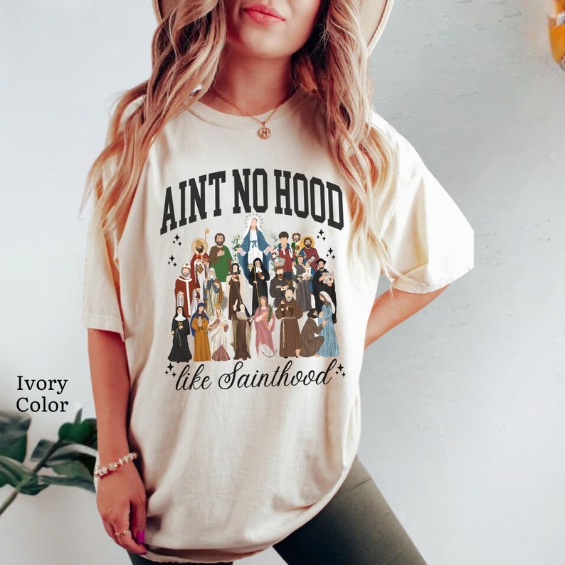 May include: A white t-shirt with a black graphic that says "Ain't No Hood Like Sainthood" and features an illustration of a group of saints.