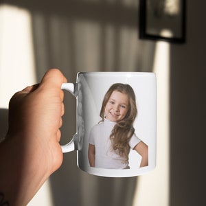 May include: White ceramic mug with a photo of a young girl with long brown hair smiling. The girl is wearing a white turtleneck shirt.