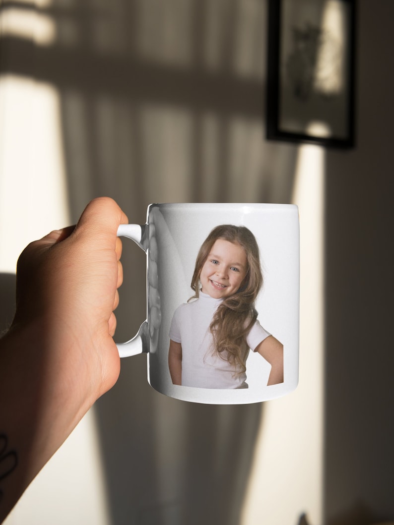 May include: White ceramic mug with a photo of a young girl with long brown hair smiling. The girl is wearing a white turtleneck shirt.