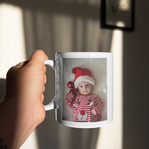 May include: White ceramic mug with a photo of a baby wearing a red Santa hat and a red and white striped onesie. The baby is holding red ornaments and is surrounded by white fluffy material.