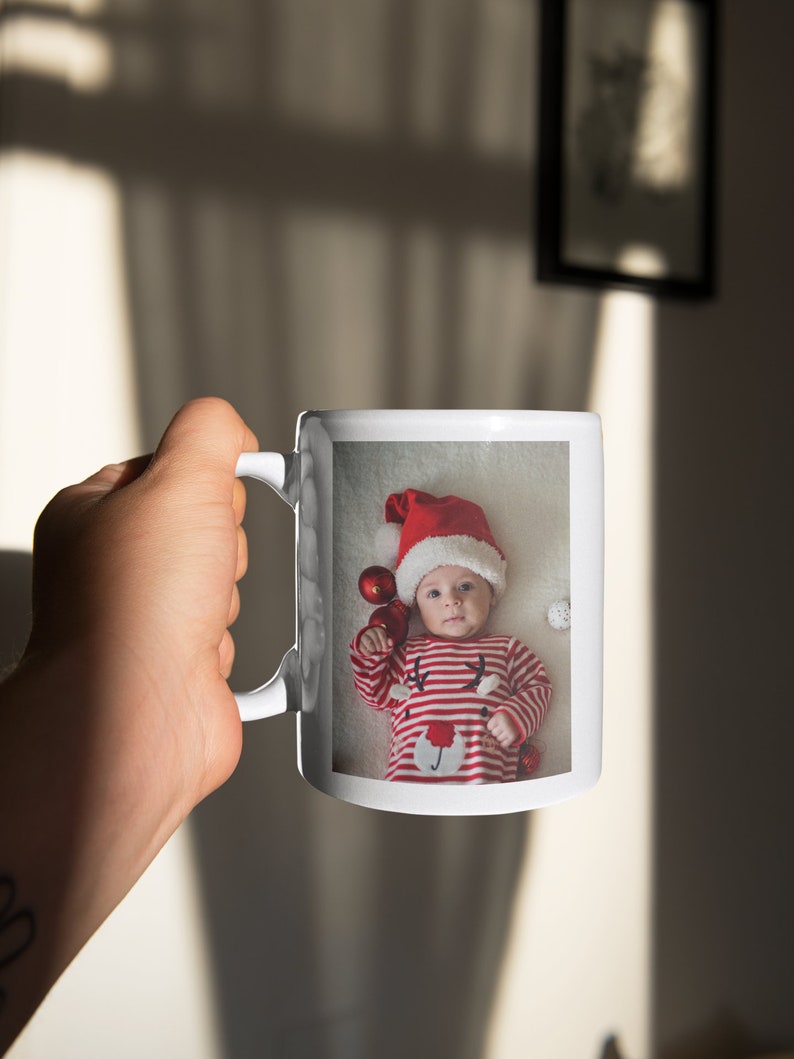 May include: White ceramic mug with a photo of a baby wearing a red Santa hat and a red and white striped onesie. The baby is holding red ornaments and is surrounded by white fluffy material.