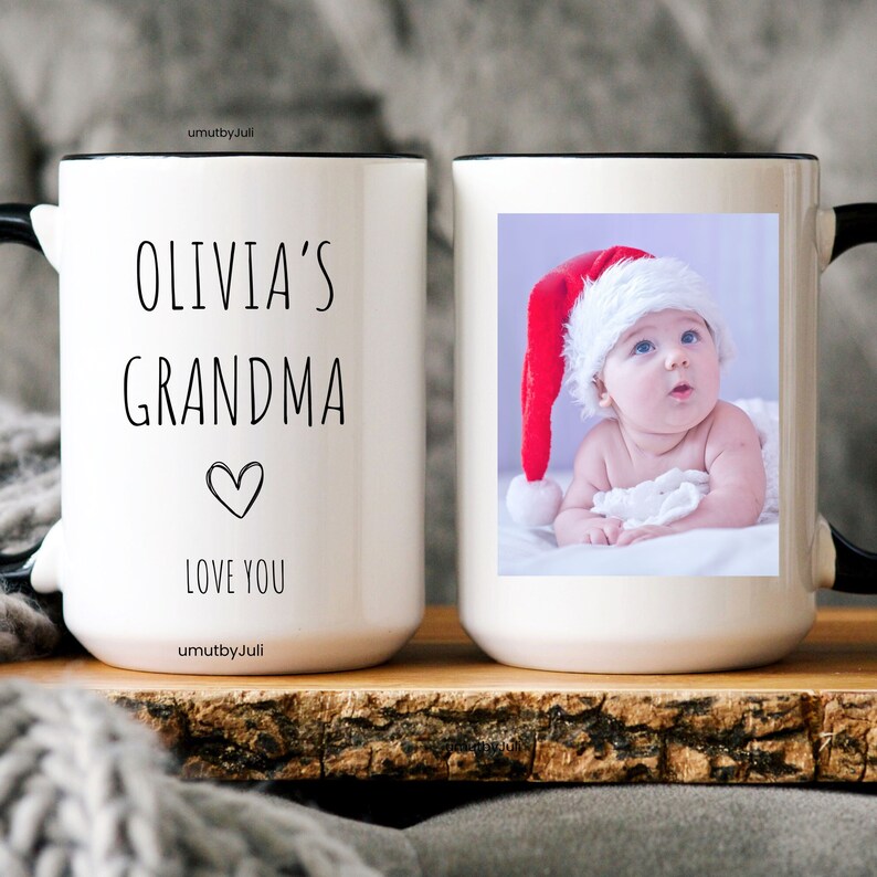 May include: White ceramic mug with a black rim and handle. The mug has a photo of a baby wearing a red Santa hat on one side and the text "Olivia's Grandma Love You" on the other side.