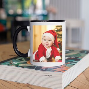 May include: White ceramic mug with a black handle and a black rim. The mug features a printed image of a baby dressed in a red Santa suit, crawling towards a Christmas tree.