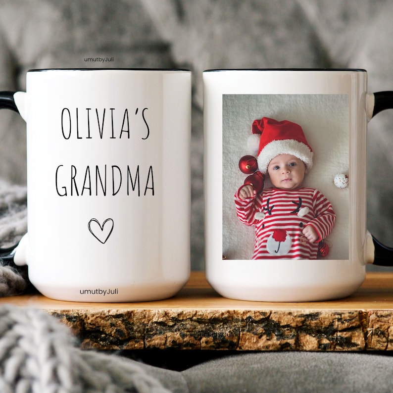 May include: White ceramic mug with a black rim and handle. The mug has a photo of a baby wearing a red and white striped outfit and a Santa hat. The text "Olivia's Grandma" is printed on the mug.