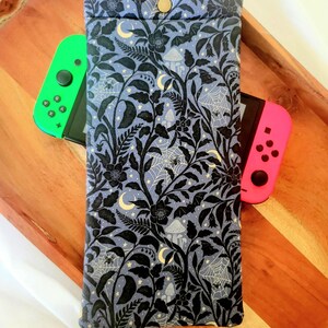May include: A blue and black patterned case for a gaming console, featuring a floral and botanical design with moons, mushrooms, and spiderwebs. The case is closed with a snap. A green and pink controller are visible.