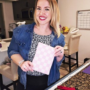 Puede incluir: Una mujer sostiene una bolsa de tela a cuadros rosa y blanca. La bolsa est&aacute; hecha de una tela suave y acolchada. La mujer lleva una chaqueta vaquera azul y una camisa de flores blancas y negras. Est&aacute; de pie en una habitaci&oacute;n con una mesa y una silla.