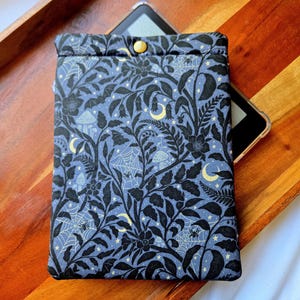 May include: A blue and black patterned e-reader sleeve with a button closure. The fabric features a botanical design with bats, mushrooms, spiderwebs, and crescent moons. The sleeve is designed to hold an e-reader.