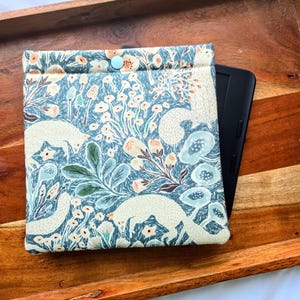 以下が含まれることがあります： 薄い青色のボタン留め具が付いた、青とクリーム色の花柄の電子書籍リーダーケース。ケースは、幻想的な花と動物のプリントでデザインされています。