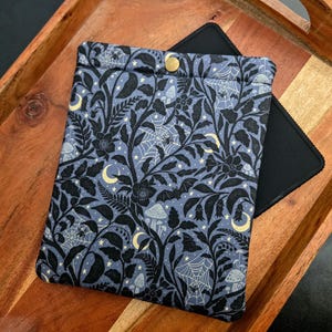 May include: A blue and black patterned tablet sleeve with a gold snap closure. The fabric features a repeating design of bats, spiderwebs, mushrooms, and crescent moons. The sleeve is resting on a wooden tray, with a black tablet partially visible.
