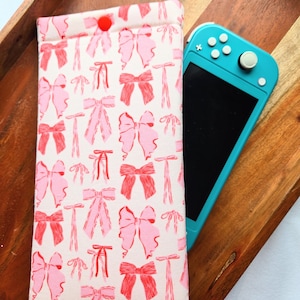 May include: A white fabric case with a pink and red bow pattern. The case has a red button closure and is designed to hold a Nintendo Switch.