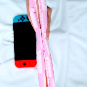 Puede incluir: Una bolsa de tela a cuadros rosa y blanca con forro blanco. La bolsa est&aacute; doblada por la mitad y sostenida por una mano.