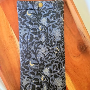 May include: A rectangular, blue fabric case with a button closure. The case features a repeating pattern of black floral and botanical elements, including flowers, leaves, mushrooms, spiderwebs, bats, moons, and stars. The case is resting on a wooden tray.