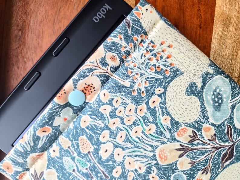 以下が含まれることがあります： 青い花柄のケースに入った黒いKobo電子書籍リーダー。ケースには薄い青色のボタンが付いている。