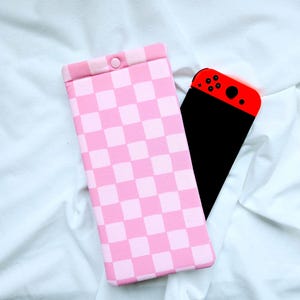 May include: A pink and white checkered case for a Nintendo Switch console. The case has a snap closure.