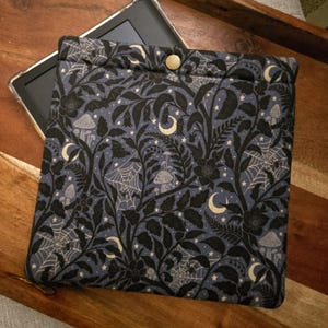 May include: A padded e-reader case with a button closure. The case features a dark blue background with a repeating pattern of black leaves, flowers, bats, spiderwebs, mushrooms, moons, and stars. The case is designed to hold an e-reader.