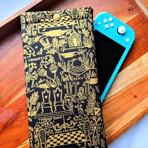 May include: Black and gold fabric case with a whimsical Alice in Wonderland design featuring illustrations of playing cards, a teapot, a throne, and the words "Mad Tea Party", "Croquet", "The Beach", and "Duchess's House".