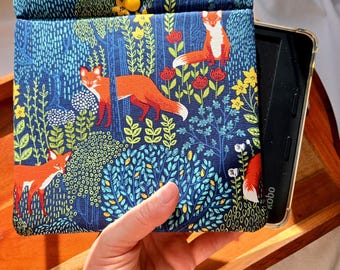 May include: A blue and green fabric e-reader case with a fox print. The case has a yellow button and is shown with a Kobo e-reader.