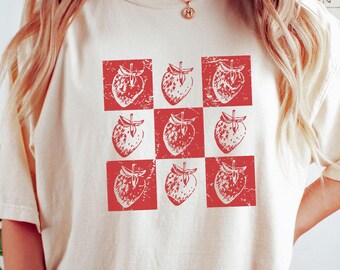 May include: A white t-shirt with a red checkered pattern featuring a repeated image of a strawberry.