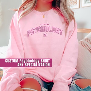 May include: A light pink sweatshirt with the words "CLINICAL PSYCHOLOGY" in purple lettering. Below the text is a purple psi symbol. The bottom of the image has a banner that reads "CUSTOM PSYCHOLOGY SHIRT ANY SPECIALIZATION".