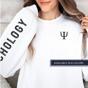 May include: White sweatshirt with the word "Psychology" printed on the left sleeve and a psychology symbol on the front. The text "AVAILABLE IN 6 COLORS" is printed on a blue rectangle at the bottom of the image.