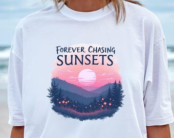 May include: White t-shirt with a graphic of a sunset over mountains. The design features a pink and purple sky, a large sun, and the text "FOREVER CHASING SUNSETS" in navy blue.