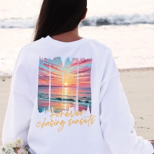 Może przedstawiać: Biała bluza z nadrukiem zachodu słońca na plecach. Grafika przedstawia kolorowy zach&oacute;d słońca nad oceanem z napisem "forever chasing sunsets" kursywą. Bluza jest noszona na plaży.