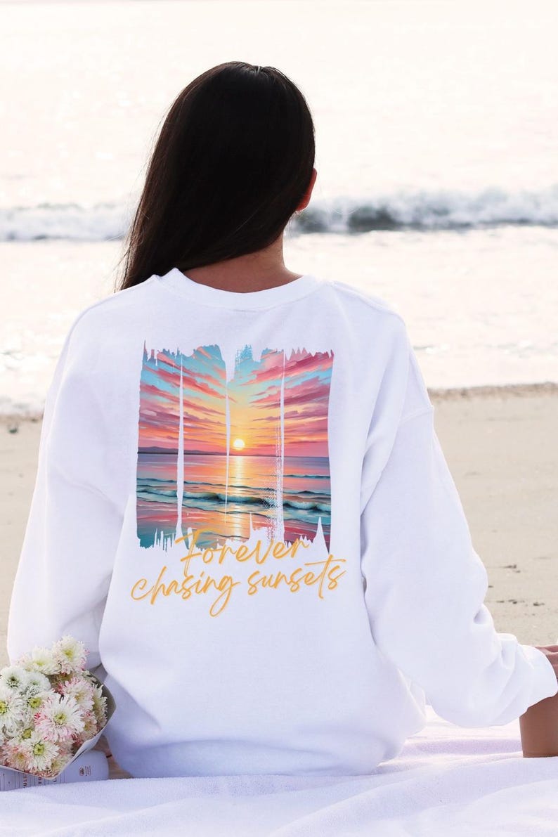 Może przedstawiać: Biała bluza z nadrukiem zachodu słońca na plecach. Grafika przedstawia kolorowy zach&oacute;d słońca nad oceanem z napisem "forever chasing sunsets" kursywą. Bluza jest noszona na plaży.