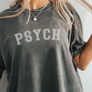 May include: A dark gray t-shirt with the word "PSYCH" printed in white on the front. The shirt is made of a soft, comfortable fabric and has a relaxed fit.